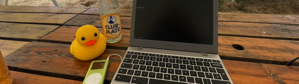 A laptop, a classic iPod Nano, an empty bottle of Club Mate, and a rubber duck all sit on a wooden table.