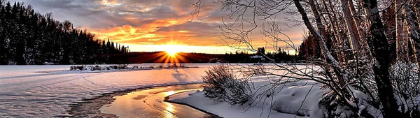 A snowy field is lit by a sun setting. There is a semi-frozen river running,