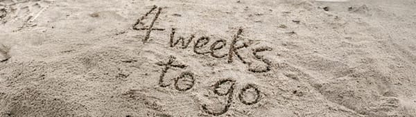 The words 'four weeks to go' are written in sand on a beach.